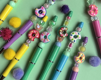 May include: A collection of colorful pens with decorative donut-shaped toppers and beaded accents. The pens feature various colors, including yellow, green, purple, and pink, with silver metal accents. The background is a light green.