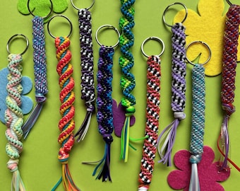 May include: A collection of colorful handmade keychains with metal rings and decorative tassels. The keychains feature various patterns and color combinations, including red, blue, green, and purple. They are arranged on a green background with felt flower accents.