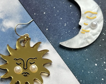 May include: Gold sun and white moon earrings. The sun earring has a smiling face with pearl accents. The moon earring has a face design with yellow accents and a pearl. The earrings are on gold hooks.