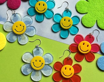 May include: A collection of flower-shaped earrings in various colors, including blue, red, and clear, each featuring a yellow smiley face center. The earrings have silver hooks and are displayed on a green and gray background.