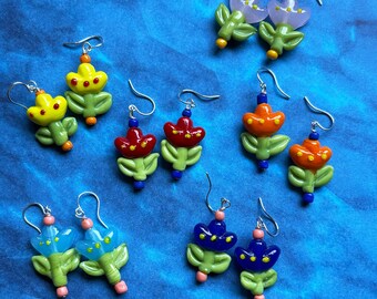 May include: A collection of six pairs of glass flower earrings featuring various colors including yellow, red, orange, light blue, dark blue, and lavender. Each earring has a green glass stem and leaf base with silver-toned metal hooks.