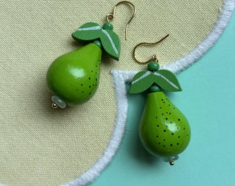 May include: A pair of handmade green pear-shaped earrings with black speckles and gold-colored hooks. The earrings have green leaf accents with white details and a small bead at the base. The earrings are on a yellow background.