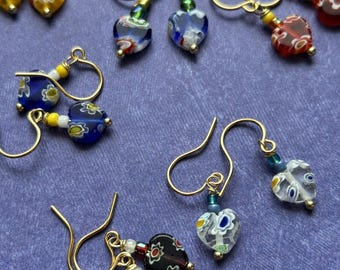 May include: A collection of handmade heart-shaped earrings with gold-colored hooks. The earrings feature colorful glass beads in various designs, including floral and abstract patterns. The color palette includes red, blue, yellow, and white, set against a purple background.