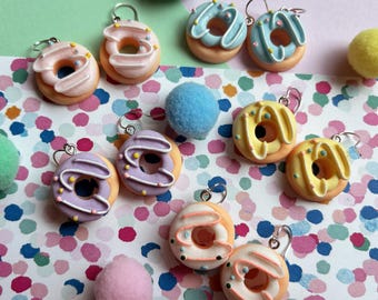 May include: A collection of colorful donut earrings with various frosting colors, including pink, blue, yellow, and purple. Each earring has a small silver hook. The donuts are arranged on a colorful polka dot background.