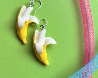 May include: A pair of novelty earrings featuring a yellow banana design with white exposed fruit. The earrings have silver-colored fish hook ear wires. The background is a solid green with pink and blue tubing.
