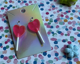 May include: A pair of heart-shaped lollipop earrings in bright pink, with white sticks, displayed on a silver card. A small green frog and a light blue flower are also visible on a colorful polka dot background. The earrings are a fun, playful accessory.
