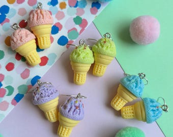 May include: A collection of colorful ice cream cone earrings. The earrings feature yellow cones and scoops of pink, green, purple, and blue ice cream. The background includes colorful confetti and pom-poms.