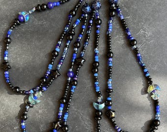 May include: Several beaded necklaces in black, blue, and purple. The necklaces feature a variety of beads, including small round beads, star-shaped beads, and moon-shaped beads. The necklaces are strung on black cord.