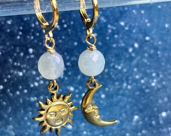 May include: A pair of gold-toned hoop earrings featuring a dangling white, faceted, iridescent bead on each. One earring displays a gold sun charm with a face, while the other features a gold crescent moon charm with a face, both suspended from wire.