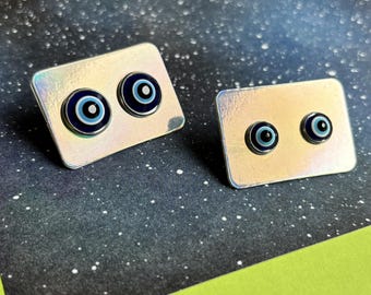 May include: Two pairs of silver-toned rectangular stud earrings, each featuring two blue and black evil eye designs. The earrings have a shiny, iridescent finish, and are set against a dark, speckled background.