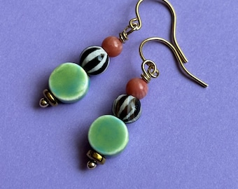 May include: A pair of handmade earrings with gold-colored ear wires. Each earring features a light green disc bead, a black and white striped bead, a coral-colored bead, and gold-colored accents. The earrings are displayed on a purple background.