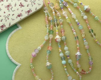 May include: A collection of phone charms featuring pastel-colored beads in various shapes, including spheres, stars, and flowers. The charms are strung on thin pink cords and include translucent, opaque, and glittery finishes in shades of pink, green, blue, and yellow.