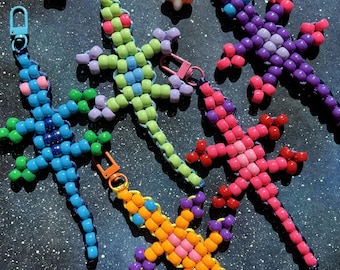 May include: A collection of beaded lizard keychains in various colors, including blue, green, pink, purple, and orange. Each lizard has a different color scheme and a metal clasp for attaching to keys or bags. The background is a dark blue with star-like accents.