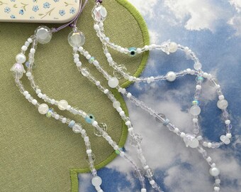 May include: A phone charm strap featuring a variety of white, clear, and iridescent beads. The design includes small flower-shaped beads, round pearls, and several blue and white evil eye charms. The strap is attached to a cream phone case.