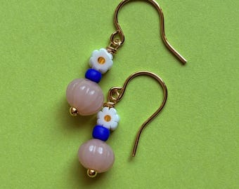 May include: A pair of gold-colored dangle earrings. Each earring features a light pink, round bead, a white daisy bead, and a dark blue bead. The earrings have gold-colored ear wires and small gold-colored accents.