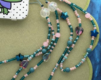 May include: A phone charm strap featuring a mix of teal, light pink, and clear crackle beads, accented with dark blue star-shaped beads and iridescent faceted charms. The strap is attached to a white phone case with a green alligator sticker.