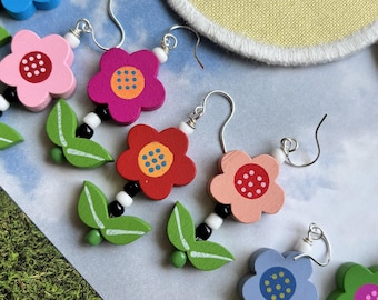 May include: Wooden flower earrings in various colors, including pink, red, and blue, with white and black bead accents. The earrings feature silver-colored hooks and green leaf charms. The flowers have a dotted center design.