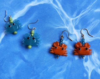 May include: Four pairs of crab-shaped earrings. Two pairs are blue, and two are orange. Each earring has a silver hook and small beads in green and blue. The earrings are set against a blue background.