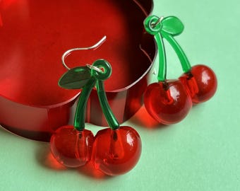May include: A pair of cherry-shaped earrings with red translucent orbs and green stems and leaves. The earrings are set against a red heart-shaped background and a light green surface.