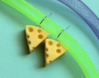 May include: A pair of novelty earrings featuring yellow cheese wedge charms with holes, suspended from silver hoops. The earrings are set against a light blue background, with neon green and purple mesh tubing in the background.