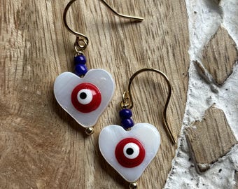 May include: A pair of gold-tone hook earrings featuring white heart-shaped charms. Each charm displays a central red, white, and black evil eye motif. A small dark blue bead sits above each heart, connecting the charm to the gold-tone wire hook.
