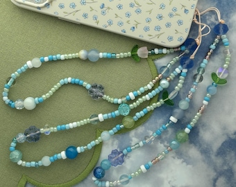 May include: A phone charm strap featuring a variety of blue, green, and white beads in different shapes and sizes, including small flower-shaped beads and translucent spheres, attached to a light-colored phone case with a blue floral pattern.