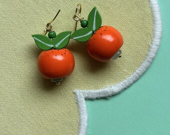 May include: A pair of orange fruit earrings with green leaves and gold-colored hooks. The earrings are made of wood and have a glossy finish. The leaves have white stripes. The earrings are on a yellow background.