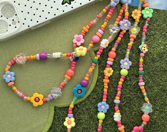 May include: A phone charm strap featuring a colorful array of plastic beads in shades of pink, orange, yellow, green, blue, and purple. The design includes various flower-shaped beads, round beads, and a small yellow smiley face bead.
