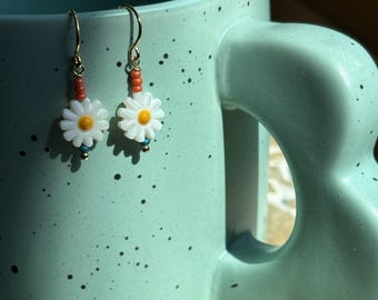 May include: A pair of daisy earrings with gold-colored hooks. Each earring features a white daisy with a yellow center, accented by orange and turquoise beads. The earrings are displayed against a light blue background.