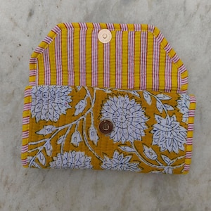 Pode incluir: Uma carteira de tecido acolchoado com base amarelo-mostarda, estampa floral branca no exterior e um padrão de listras verticais amarelas e rosas na aba interna. A carteira inclui um fecho de botão de pressão metálico circular.