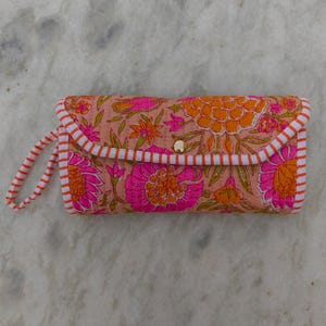 Può includere: Una pochette rettangolare trapuntata con sfondo color pesca caratterizzata da un vivace motivo floreale nei toni del rosa acceso e dell'arancione. L'accessorio include un cinturino da polso abbinato e un bordo a righe bianche e arancioni.