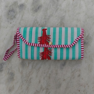 Può includere: Una pochette trapuntata rettangolare con strisce verticali color verde acqua e bianco. La patta è rifinita con un bordo a righe rosse e bianche e una chiusura a scatto dorata. Due illustrazioni di aragoste rosse sono centrate sul davanti, con un cinturino da polso a righe rosse e bianche abbinato sul lato.