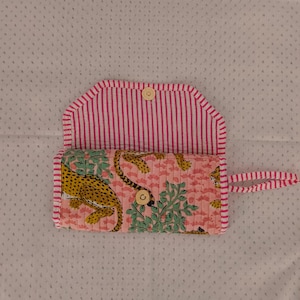 Pode incluir: Uma bolsa de tecido acolchoado com estampa de leopardo em fundo rosa e folhagem verde. O interior e as bordas são forrados com listras verticais rosa e brancas. Inclui um fecho de botão de pressão dourado e uma alça de tecido.