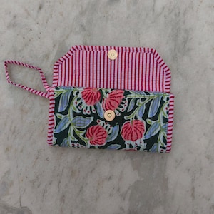 Può includere: Un portafoglio in tessuto trapuntato con base nera e motivo floreale rosa e azzurro. La patta interna e il cinturino da polso presentano un design a righe rosse e bianche. Una chiusura magnetica color oro mantiene il portafoglio chiuso.