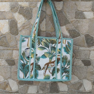 Puede incluir: Un bolso tote acolchado con base blanca y un patrón repetitivo de hojas tropicales verdes y leopardos manchados de color marrón. El bolso incluye dos asas largas para el hombro y está rematado con un borde de tela a rayas verdes y blancas.