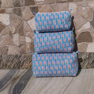 Può includere: Tre trousse per cosmetici blu trapuntate impilate. Ogni borsa è decorata con un motivo floreale rosa ripetuto. Le borse hanno una chiusura a cerniera e piccoli anelli di tessuto blu su ciascuna estremità.