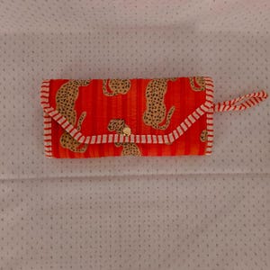 Peut inclure: Un portefeuille rectangulaire en tissu matelassé orange vif avec des rayures verticales et un motif léopard beige répétitif. Le portefeuille présente un rabat avec une fermeture à pression dorée et une dragonne en tissu rayé rouge et blanc.