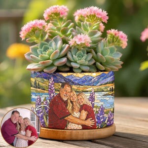 May include: A succulent plant with light green leaves and pink flowers sits in a cylindrical planter. The planter features a mosaic-style print of a family of three embracing in front of a landscape with purple lupine flowers and blue mountains.