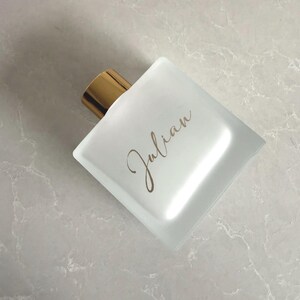 May include: A square, frosted glass perfume bottle with a gold-coloured cap. The word "Julian" is written in a cursive font on the front of the bottle. The bottle is on a light grey, marble-like surface.