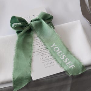 May include: A sage green ribbon tied in a bow, with frayed edges, rests on a white napkin. The word "YOUSSEF" is printed in white on one of the ribbon tails. A menu is visible beneath the ribbon and napkin.
