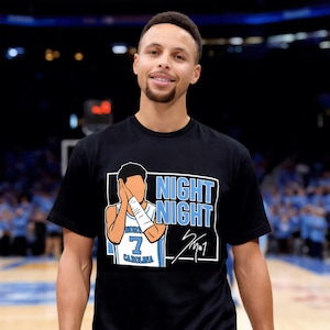 Può includere: T-shirt nera con una grafica a cartoni animati di un giocatore di basket e le parole "NIGHT NIGHT" in azzurro e bianco. Il giocatore indossa una maglia con il numero 7 e "CAROLINA". La maglietta ha anche una firma.