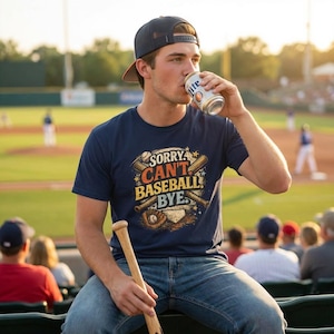 May include: A navy blue t-shirt with the text "Sorry Can't Baseball Bye" in a baseball-themed design. The shirt is worn by a person at a baseball game, holding a baseball bat and drinking from a can of beer.