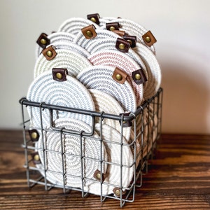 May include: A wire basket filled with a stack of round, woven cotton pot holders with leather handles. The pot holders are in shades of beige, gray, and pink.