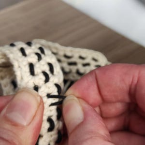 Könnte beinhalten: Ein gehäkeltes Band mit einer cremefarbenen Garnbasis und schwarzen Garnakzenten, die in einem sich wiederholenden Muster durch die Maschen gewebt sind. Die Textur ist dick und strukturiert und zeigt die komplexe Schlaufenkonstruktion des Textils.
