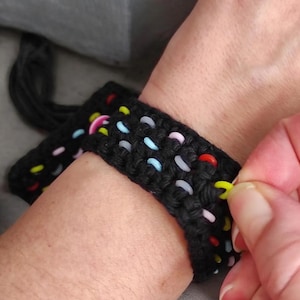 Op de afbeelding: Een zwart gehaakt armband met kleurrijke kralen in rood, geel, roze en blauw. De armband wordt vastgemaakt met een gele sluiting. De armband zit om een pols.
