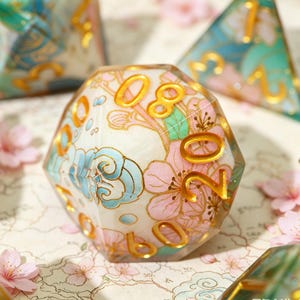 May include: A multi-sided die with a floral design in pink, blue, and gold. The die features gold numbers and is set against a background with similar floral patterns. The overall aesthetic is delicate and ornate.
