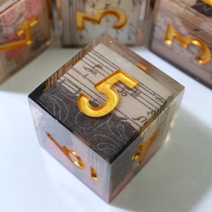 May include: A clear resin cube containing a piece of paper with black musical notation and a dark patterned background. A raised, metallic gold-colored number five is embedded on the top surface, with other gold numbers visible on the sides.