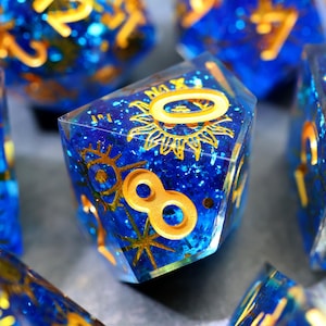 May include: Close-up of a set of polyhedral dice. The dice are a deep blue with gold-coloured numbers and celestial designs. The dice are made of a clear resin with glitter and embedded gold accents. The number eight is visible on one die.