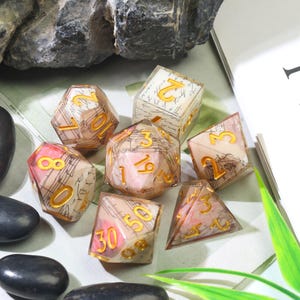 May include: A set of seven polyhedral dice made of clear resin with embedded paper featuring musical notation and script. Each die has gold-colored numbers. The set includes a d20, d12, d10, d8, d6, d4, and a percentile die, all displaying a vintage aesthetic.