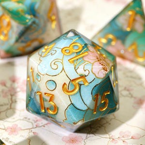 Może przedstawiać: Zestaw wielościennych kości do gry z kwiatowym wzorem. Kości są w odcieniach niebieskiego, białego i r&oacute;żowego ze złotymi numerami. Idealne do gier RPG.